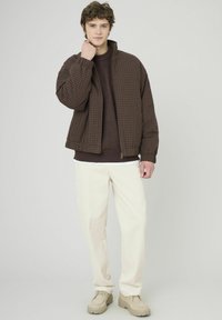 Doudoune à carreaux marron avec fermeture éclair, portée sur un pull foncé. Pantalon en velours côtelé crème et chaussures beiges épaisses complètent la tenue.