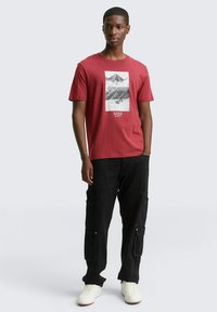 Rode katoenen T-shirt met grafische print van bergen en tekst; gecombineerd met zwarte cargobroek met meerdere zakken; witte sneakers.