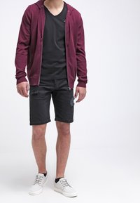 Burgunderfärgad zip-up hoodie över en svart v-ringad t-shirt, tillsammans med slitna svarta shorts och vita sneakers med grå snören.