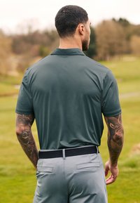 Polo shirt homme vert foncé avec un motif texturé, associé à un pantalon gris clair avec une taille noire. La vue arrière montre des bras tatoués.