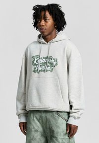 Felpa grigia chiara con un grande design grafico di testo verde e bianco. Dotata di tasca a marsupio e cappuccio regolabile con cordino.