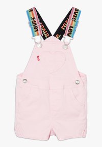 Salopette di jeans rosa con hardware argentato e dettagli cuciti a forma di cuore. Spallacci regolabili neri e a righe arcobaleno.