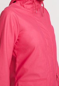 Vattentät rosa jacka med huva, med en slät yta, framdragkedja och sidofickor. Skräddarsydd passform med kontrasterande sömmar.