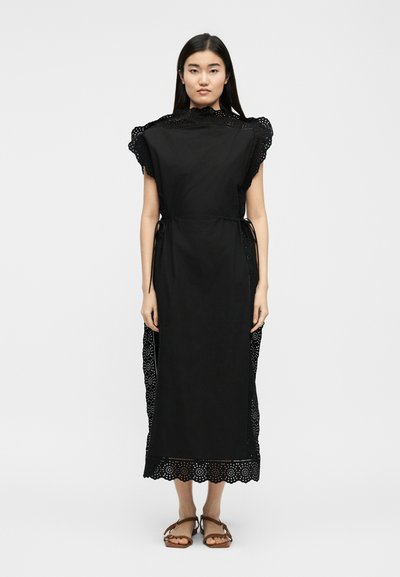 Γυναίκα στέκεται φορώντας μαύρο midi φόρεμα με διακοσμητική δαντέλα eyelet και κοντά μανίκια, συνοδευόμενο από καφέ επίπεδα σανδάλια, σε απλό λευκό φόντο.