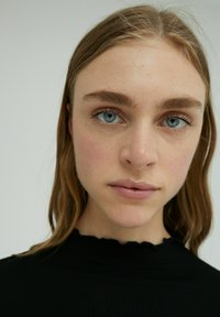 Nahaufnahme einer Person mit blauen Augen und hellbraunem Haar. Sie trägt ein schwarzes, geripptes Oberteil mit einem gewellten Ausschnitt. Glatte Haut, natürliches Make-up.