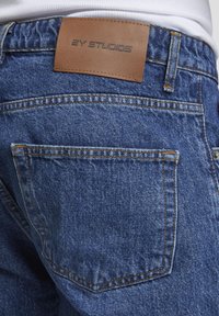 Buzunar din spate al blugilor din denim albastru, cu cusături portocalii și o pată de piele maro etichetată „2Y STUDIOS” pe talie.