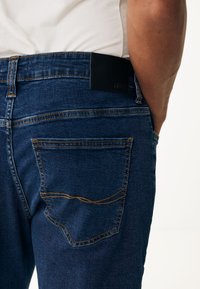 Mörkblå denimjeans med en slät yta, med bruna sömmar, två bakfickor och en svart läderrem på midjan.