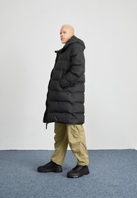 Zwarte puffer lange jas met een capuchon, ritssluiting en zijzakken, gecombineerd met beige wijde broek en zwarte platform sneakers.