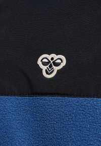 Fleecejack dat een blauw onderpaneel heeft met een gestructureerde afwerking en een zwart bovenste gedeelte, versierd met een wit geborduurd bijenlogo.