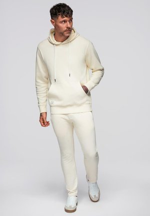 Mand med krøllet hår iført cremefarvet hættetrøje, matchende sweatpants og hvide sneakers, ser mod venstre med den ene hånd i lommen.