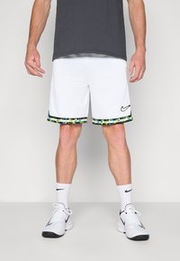 Baltas sporta šortus ar krāsainu rakstu apmalēm apakšējā daļā, ar melnu Nike logotipu, kombinēti ar baltām sporta kurpēm un zeķēm.