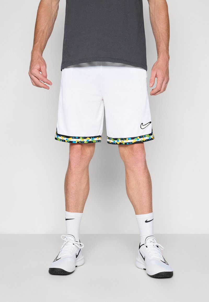 Baltas sporta šortus ar krāsainu rakstu apmalēm apakšējā daļā, ar melnu Nike logotipu, kombinēti ar baltām sporta kurpēm un zeķēm.