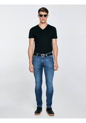 Férfi fekete V-nyakú pólóban, kék farmerben, fekete övvel, fekete tornacipőben és sötét napszemüvegben, egyszínű háttér előtt áll.