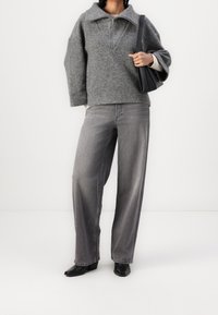 Suéter gris de gran tamaño con cuello de cremallera, combinado con jeans grises de pierna ancha. Bolso de hombro negro y botas negras de tacón completan el conjunto.