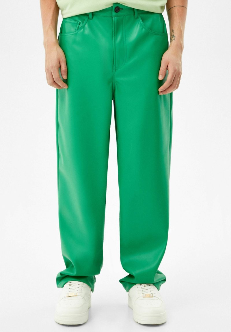 Homme portant un pantalon ample vert vif et des baskets blanches, debout devant un fond blanc uni.