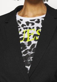 Černý blazer s klopou, nosený přes bílý tričko s černobílým a neonově žlutým leopardím vzorem.