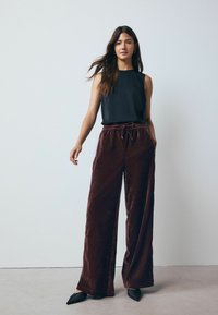 Schwarzes ärmelloses Oberteil mit Rundhalsausschnitt; burgunderrote weite Samthose mit Kordelzugbund; schwarze spitze Ballerinas.