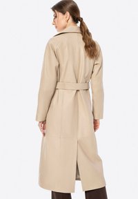 Trench coat in pelle beige con vita elasticizzata, maniche lunghe e taglio a metà lunghezza. Presenta una fessura sul retro e un colletto strutturato.