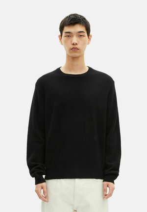 Jeune homme portant un pull noir à manches longues et un pantalon clair, se tenant devant un fond blanc uni.