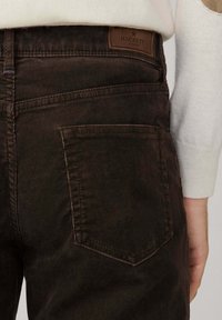Vista trasera de una persona que lleva pantalones de pana marrón oscuro con una etiqueta de cuero Hackett London encima del bolsillo trasero derecho y un suéter blanco.