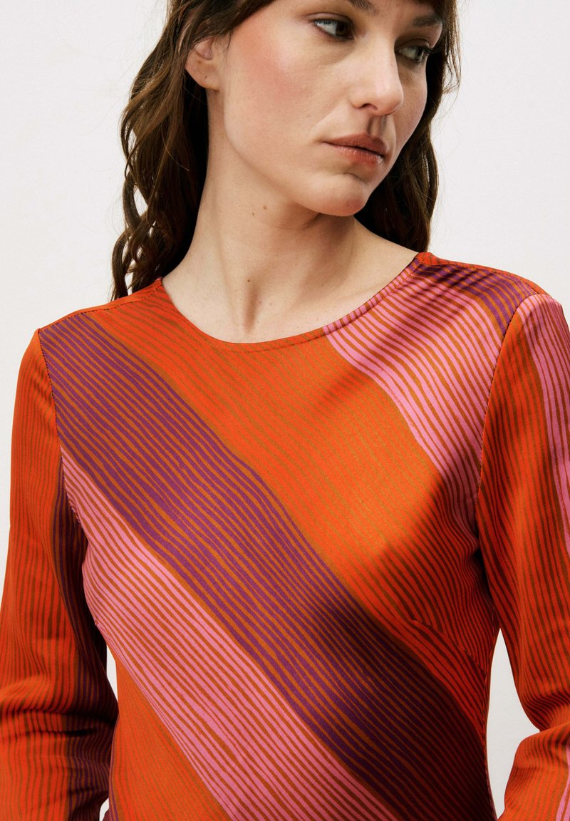 Femme aux cheveux bruns ondulés portant un haut à manches longues avec des rayures diagonales rouges, violettes et roses, regardant vers sa droite sur un fond uni.