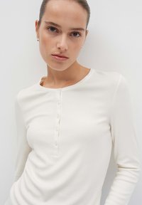Jonge vrouw met lichte huid en opgestoken haar, gekleed in een witte geribde longsleeve henley shirt met knopen, staand tegen een effen muur.