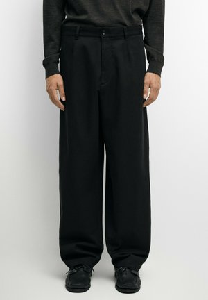 Uomo che indossa pantaloni neri a gamba larga, scarpe nere e una camicia scura a manica lunga, in piedi davanti a uno sfondo semplice.