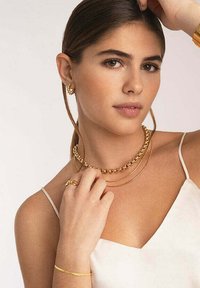I gioielli in tonalità oro includono una collana a catena a strati, orecchini a cerchio e un anello. La modella indossa un top senza maniche di colore chiaro.