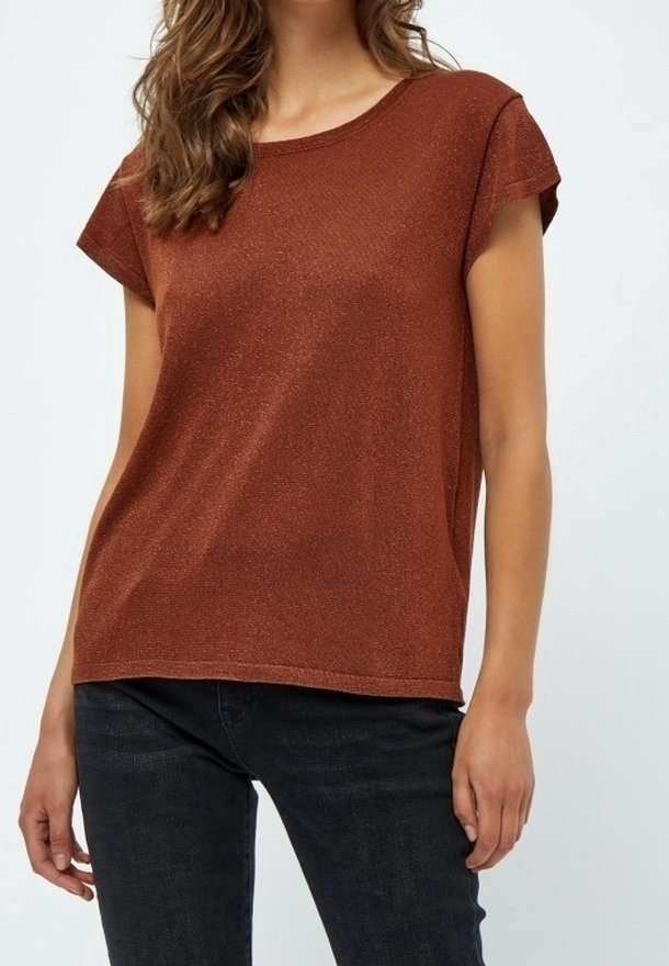 Femme portant un haut en maille rouille à manches courtes et un jean noir, debout dans une posture détendue devant un fond uni.