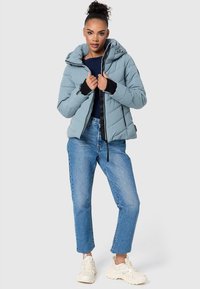 Lichtblauwe pufferjack met een capuchon, voorzien van een ritssluiting, elastische manchetten en diagonale stiksels, gecombineerd met lichtblauwe jeans en witte sneakers.