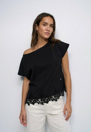 Mujer de pie contra un fondo liso, con un top negro de hombros descubiertos con ribete de encaje y pantalones blancos, mirando a la cámara.