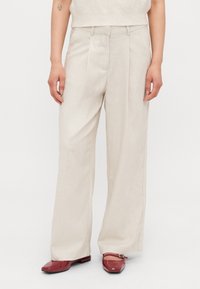 Pantalons beiges légers avec une coupe droite, un devant plissé et une coupe cintrée. Portés avec des chaussures rouges brillantes, créant un look contrasté.