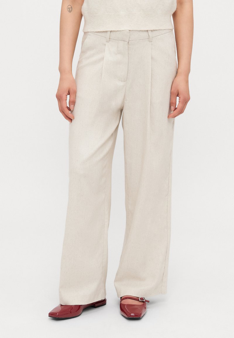 Pantalons beiges légers avec une coupe droite, un devant plissé et une coupe cintrée. Portés avec des chaussures rouges brillantes, créant un look contrasté.