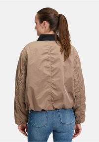Giacca bomber beige con colletto nero, caratterizzata da una superficie leggermente lucida e maniche arricciate, indossata sopra dei jeans blu.