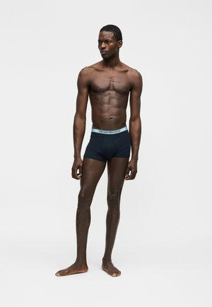 Oberkörperfreier Mann steht barfuß und trägt marineblaue Boxershorts mit einem gebrandeten hellblauen Bund vor einem schlichten hellen Hintergrund.