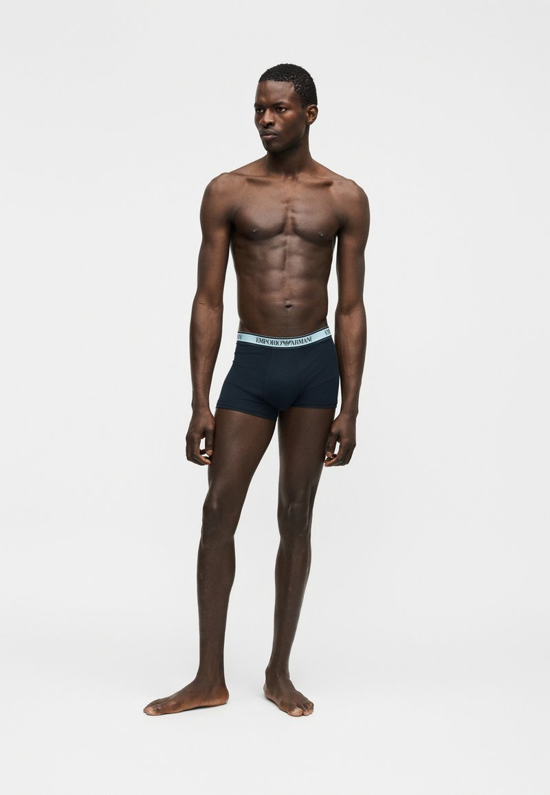 Άνδρας χωρίς μπλούζα, στεκόμενος ξυπόλητος, φοράει μπλε σκούρο boxer briefs με λουράκι μέσης σε ανοιχτό μπλε χρώμα με λογότυπο, μπροστά σε απλό ανοιχτό υπόβαθρο.