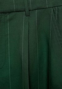 Un pantalon en tissu vert présente des rayures verticales blanches, une taille plate et des poches avant. La texture semble lisse et légèrement mate.