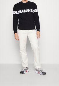 Personne portant un sweat-shirt noir avec le logo blanc "GUESS", un pantalon crème et des chaussures de sport blanches, noires et roses, debout sur un sol gris.