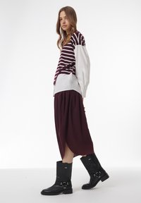 Jupe bourgogne longue, pull rayé bordeaux et blanc, et bottes noires à hauteur de genoux avec boucles et quincaillerie en argent. Style minimaliste.