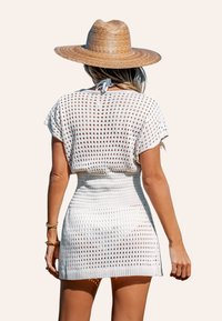 Weißes gehäkeltes Überkleid mit kurzen Ärmeln und tailliertem Schnitt, das ein strukturiertes Kabelmuster aufweist. Kombiniert mit einem Strohhut.