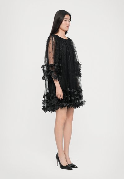 Crna haljina s prozirnim prekrivačem ukrašenim cvjetnim motivima, rukavima do tri četvrtine i višeslojnim rubom, uparena s crnim visokim potpeticama.