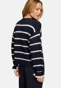 Femme portant un pull duveteux bleu marine à rayures blanches horizontales et poignets à volants, debout devant un fond clair uni.