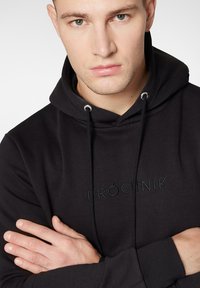 Czarna bluza z kapturem i logo z przodu, posiadająca regulowany kaptur, długie rękawy oraz ściągacze na rękawach. Wykonana z miękkiego materiału o gładkiej fakturze.