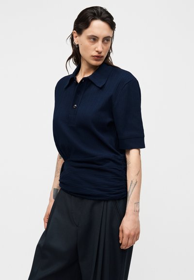 Γυναίκα με σκουρόχρωμα κυματιστά μαλλιά φοράει μπλε-ναυτικό ριμπ πικέ πόλο μπλουζάκι με κοντά μανίκια και φαρδιά μαύρα παντελόνια, στέκεται μπροστά σε έναν απλό φόντο.