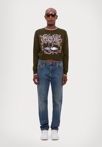 Maglione verde corto con rifiniture rosa e grafica di un cigno floreale, abbinato a jeans azzurri chiari e sneakers bianche.