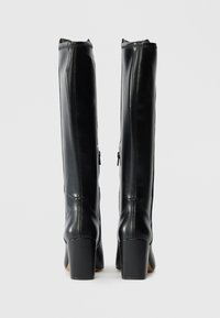 Juodos odinės kelio aukščio botai su lygia tekstūra, kvadratiniais kulnais ir šoninėmis užtrauktukais. Yra prigludusi forma ir minimalūs siuvimo detalės.