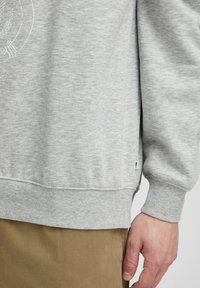 Sweatshirt gris clair avec des poignets et un ourlet côtelés, présentant un design graphique circulaire. Texture douce, avec une petite étiquette de marque sur le côté.