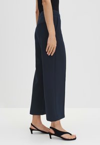 Pantalons larges bleu marine avec une taille haute, tissu texturé, poches latérales et une finition lisse, associés à des sandales à talons noires.