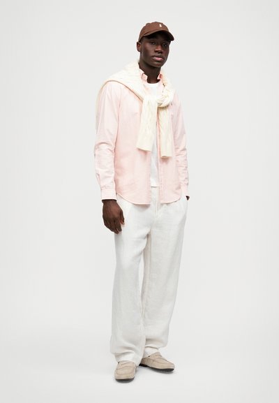 Jasnoróżowa, w paski, koszula na guziki, biała sweter narzucony na ramiona, beżowe lniane spodnie i jasnobrązowe loafersy. Brązowa czapka na głowie.