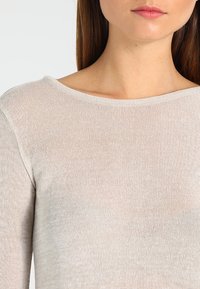 Světle béžový, lehký pletený top s širokým výstřihem do véčka a texturovanou tkaninou. Detailní záběr ukazuje jemnou, průsvitnou kvalitu materiálu.